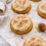 Soft cornbread cookies topped with fluffy cinnamon honey butter frosting, lightly dusted with cinnamon sugar and drizzled with honey on wrinkled parchment over a marble countertop.