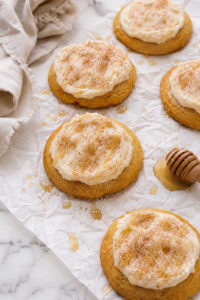 Soft cornbread cookies topped with fluffy cinnamon honey butter frosting, lightly dusted with cinnamon sugar and drizzled with honey on wrinkled parchment over a marble countertop.
