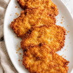 Crispy cornbread fried pork cutlets arranged on a white platter over a marble countertop with neutral linen.