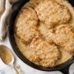 Cast iron skillet filled with crispy boneless fried pork chops smothered in creamy Southern pepper gravy on a marble countertop.