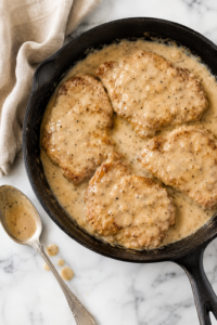 Cast iron skillet filled with crispy boneless fried pork chops smothered in creamy Southern pepper gravy on a marble countertop.
