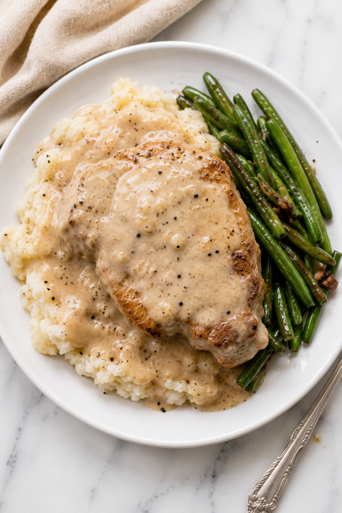 Smothered fried pork chop with creamy Southern pepper gravy served over smashed potatoes with seasoned green beans on a white plate.