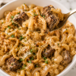 Fork lifting a bite of creamy Bloomin' Onion steak bite pasta with tender seared steak, cavatappi pasta, crispy fried onions, and rich cheese sauce from a white serving bowl.