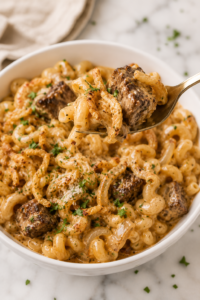 Fork lifting a bite of creamy Bloomin' Onion steak bite pasta with tender seared steak, cavatappi pasta, crispy fried onions, and rich cheese sauce from a white serving bowl.