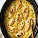 Bird's-eye view of creamy pepperoncini chicken in a black slow cooker with a rich cream sauce, whole chicken breasts, sliced pepperoncini peppers, and a wooden spoon on a marble countertop.