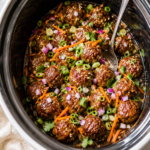 Crockpot banh mi meatballs in a slow cooker topped with fresh cilantro, shredded carrots, green onions, red onion, and sesame seeds on a marble countertop.