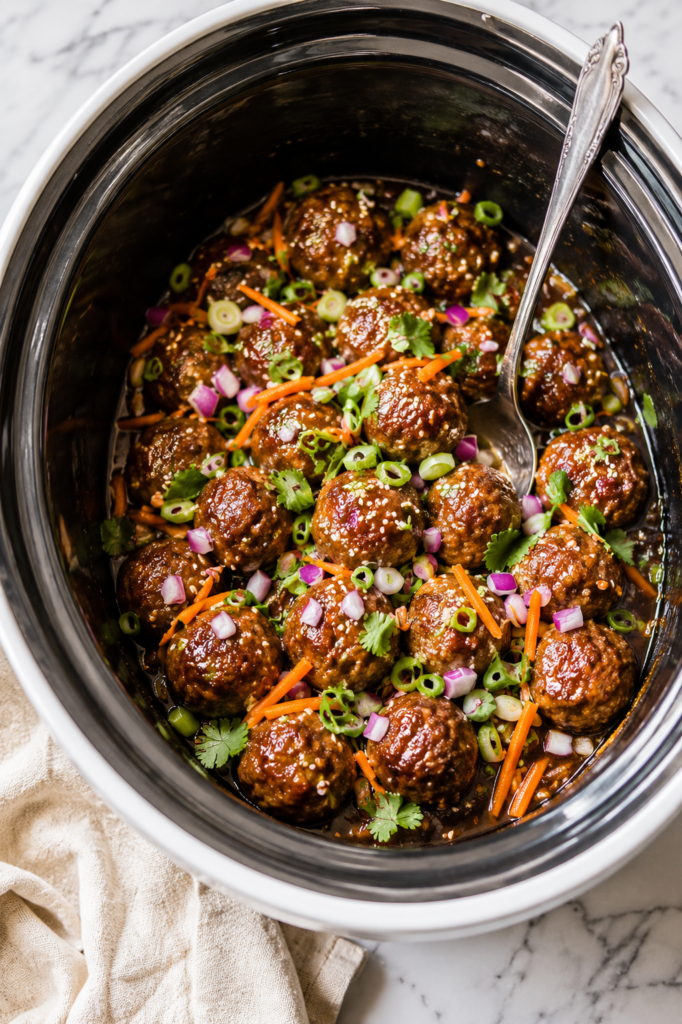 Crockpot banh mi meatballs in a slow cooker topped with fresh cilantro, shredded carrots, green onions, red onion, and sesame seeds on a marble countertop.
