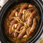 Crockpot beer brats and onions slow cooking in a black slow cooker on a white marble countertop with a neutral kitchen towel.