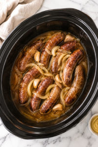 Crockpot beer brats and onions slow cooking in a black slow cooker on a white marble countertop with a neutral kitchen towel.