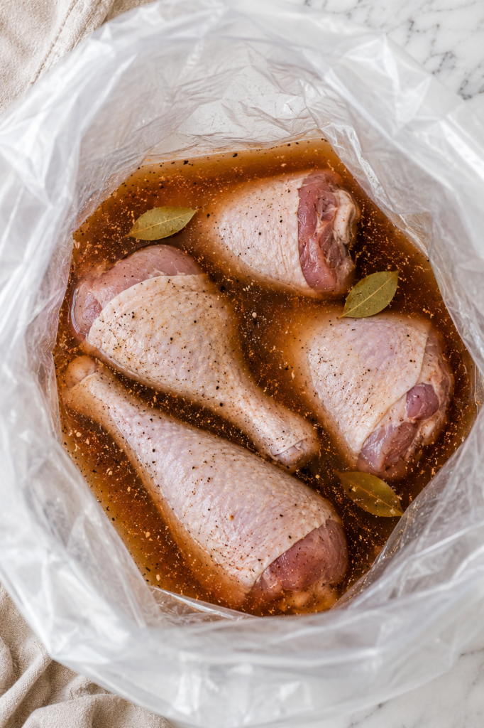 Bird's-eye view looking into a large clear food-safe brining bag filled with raw turkey legs soaking in seasoned brine with bay leaves and spices on a white marble countertop.
