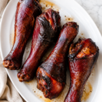 Disney World inspired smoked turkey legs served on a white platter with savory turkey drippings, set on a white marble countertop with a neutral linen cloth.