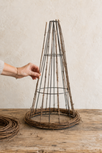 Hand attaching straight grapevine branches to a black tomato cage with curved grapevines woven around the witch hat brim.