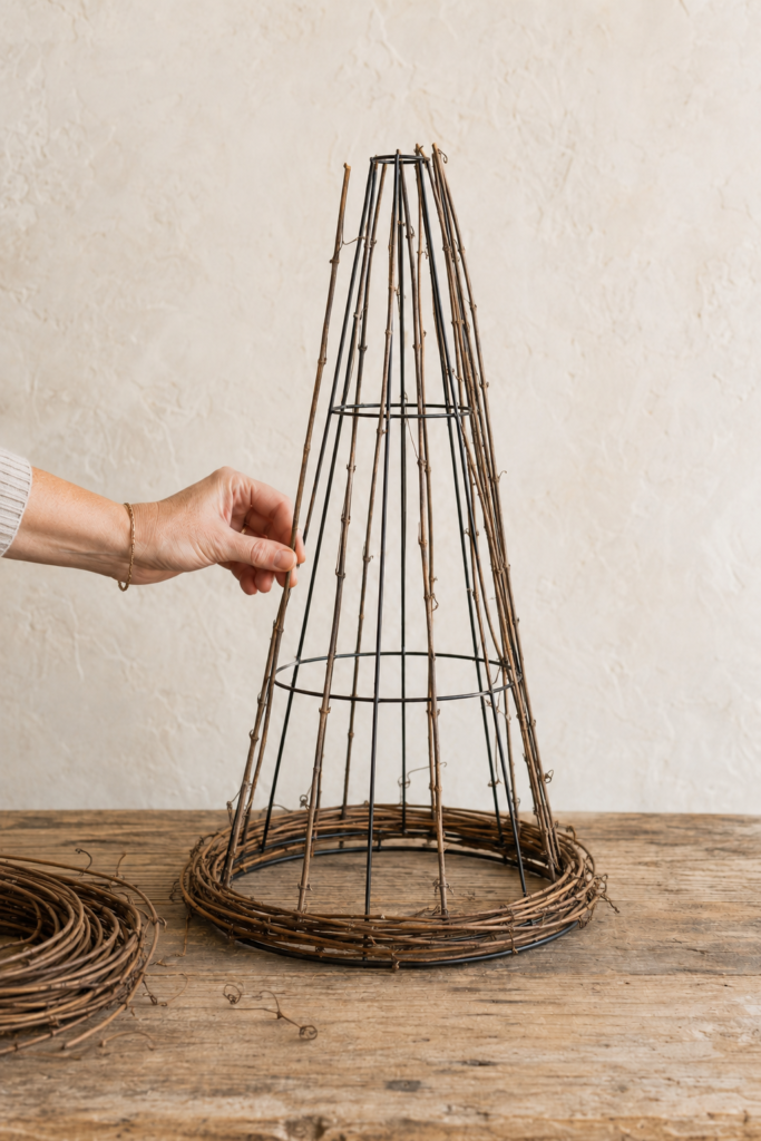 Hand attaching straight grapevine branches to a black tomato cage with curved grapevines woven around the witch hat brim.