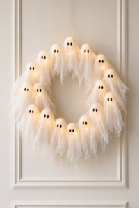 Finished DIY lighted ghost wreath with glowing cheesecloth ghosts hanging on a creamy off-white paneled wall.