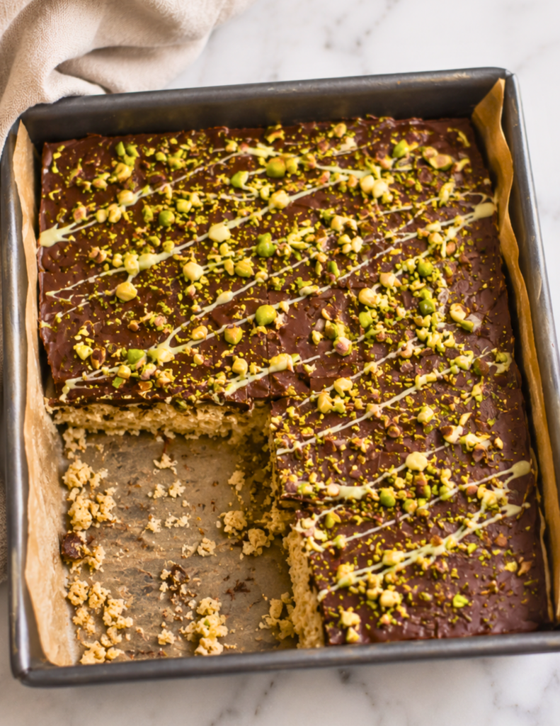 Overhead view of Dubai chocolate inspired Rice Krispie treats in a parchment-lined baking pan with a few pieces removed, topped with chocolate, chopped pistachios, and white chocolate drizzle on a marble countertop.