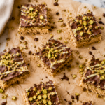 Overhead view of homemade Dubai Chocolate Inspired Rice Krispie Treats topped with chocolate, white chocolate drizzle, and chopped pistachios on crinkled parchment paper over a white marble countertop.