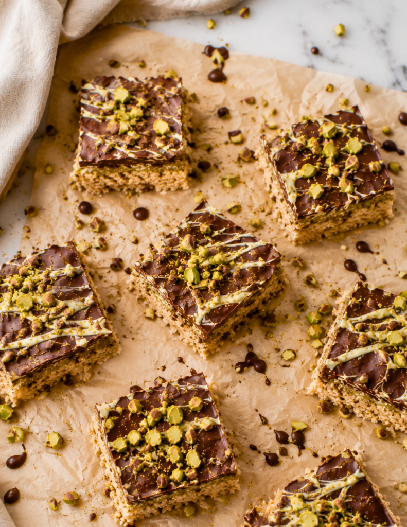 Overhead view of homemade Dubai Chocolate Inspired Rice Krispie Treats topped with chocolate, white chocolate drizzle, and chopped pistachios on crinkled parchment paper over a white marble countertop.