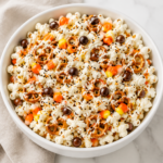 Halloween popcorn with white chocolate, pretzels, candy corn, chocolate candies, and orange and black sprinkles in a white bowl.