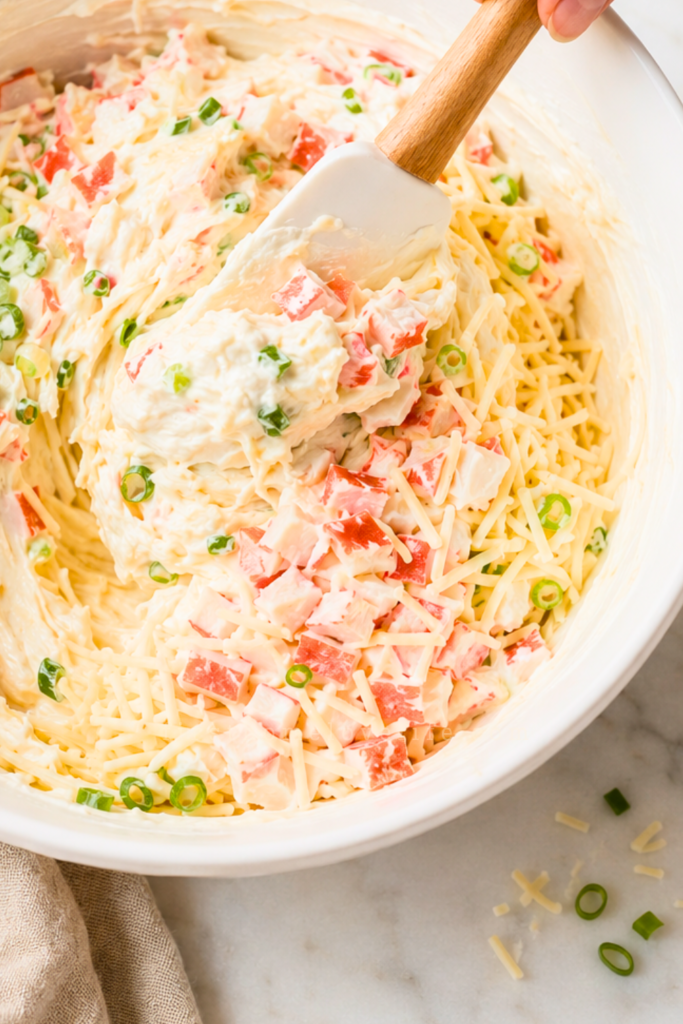 Crabmeat, mozzarella, and green onions being folded into a creamy crab rangoon dip mixture.