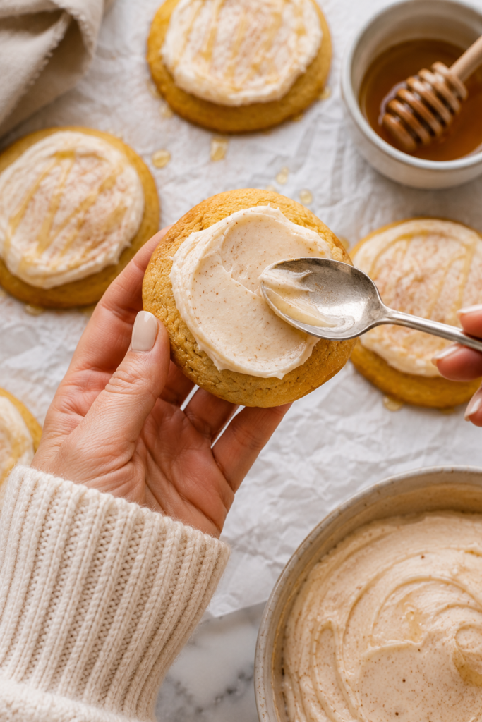 Hand holding a homemade cornbread cookie while spreading cinnamon honey butter frosting with a spoon over the top on a marble countertop.