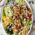 Grilled shrimp taco salad on a large white oval platter with romaine lettuce, grilled shrimp, black beans, corn, avocado, tomatoes, tortilla strips, cilantro lime dressing, and silver serving utensils on a marble countertop.
