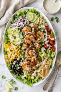 Grilled shrimp taco salad on a large white oval platter with romaine lettuce, grilled shrimp, black beans, corn, avocado, tomatoes, tortilla strips, cilantro lime dressing, and silver serving utensils on a marble countertop.