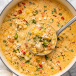 Creamy Mexican Street Corn Chicken Chowder in a white Dutch oven with a silver ladle, topped with cotija cheese, cilantro, jalapeños, and roasted corn on a marble countertop.