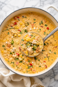 Creamy Mexican Street Corn Chicken Chowder in a white Dutch oven with a silver ladle, topped with cotija cheese, cilantro, jalapeños, and roasted corn on a marble countertop.