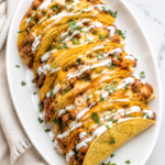 Oven baked cheesy creamy ground beef and bean tacos topped with melted cheddar, Mexican crema, and fresh cilantro on a white oval platter over a marble countertop with a neutral linen cloth.