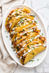 Oven baked cheesy creamy ground beef and bean tacos topped with melted cheddar, Mexican crema, and fresh cilantro on a white oval platter over a marble countertop with a neutral linen cloth.