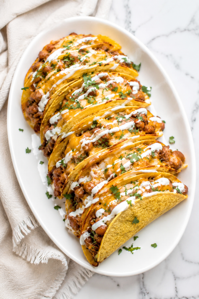 Oven baked cheesy creamy ground beef and bean tacos topped with melted cheddar, Mexican crema, and fresh cilantro on a white oval platter over a marble countertop with a neutral linen cloth.