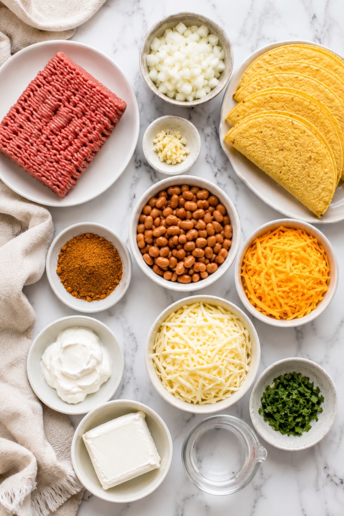 Flat lay of ingredients for oven baked cheesy creamy ground beef and bean tacos including ground beef, pinto beans, taco shells, shredded cheddar, Monterey Jack cheese, cream cheese, sour cream, onion, garlic, taco seasoning, cilantro, and water on a white marble countertop.