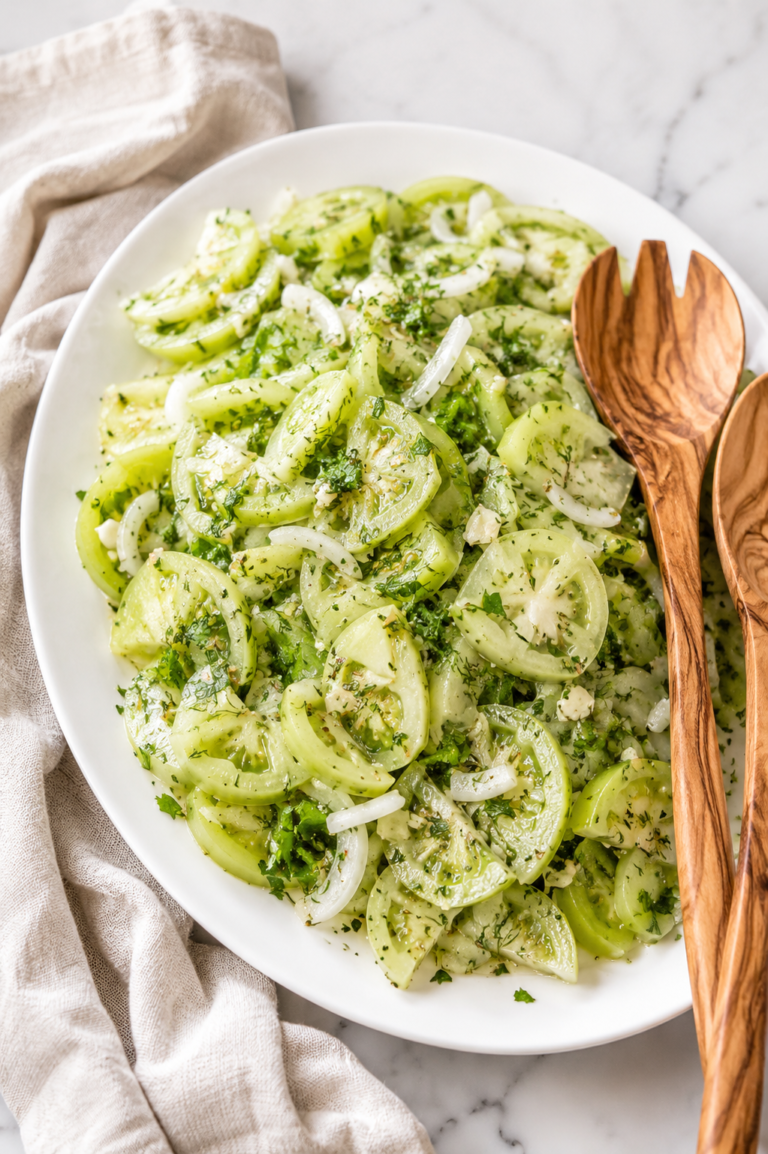 Fresh pickled green tomato salad with sliced green tomatoes, sweet onions, fresh dill, and tangy vinaigrette served on a white platter with wooden salad servers.