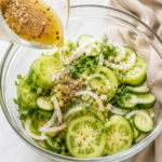 Homemade pickling brine being poured over sliced green tomatoes, cucumbers, onions, and fresh herbs in a glass mixing bowl before marinating.