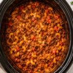 Bird's-eye view of homemade Slow Cooker Beef-A-Roni in a black crockpot with seasoned ground beef, elbow macaroni, rich tomato sauce, melted cheddar cheese, and fresh parsley on a marble countertop.