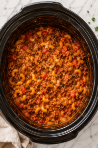 Bird's-eye view of homemade Slow Cooker Beef-A-Roni in a black crockpot with seasoned ground beef, elbow macaroni, rich tomato sauce, melted cheddar cheese, and fresh parsley on a marble countertop.
