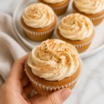 Hand holding an apple cider spiced cupcake with cinnamon cream cheese frosting and more cupcakes in the background