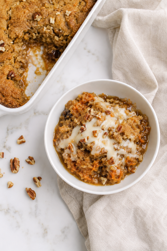 Warm carrot cake cobbler with grated carrots, pecans, gooey brown sugar sauce, and cream cheese drizzle in a white bowl.
