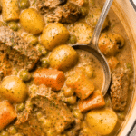 Creamy beef stew with tender beef, potatoes, carrots, mushrooms and peas cooking in a white Dutch oven with a silver ladle