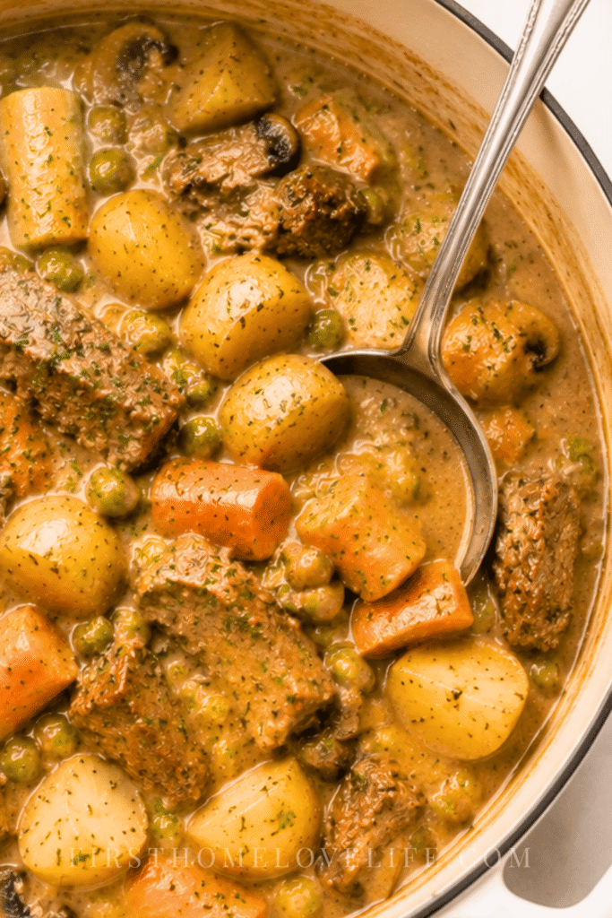 Creamy beef stew with tender beef, potatoes, carrots, mushrooms and peas cooking in a white Dutch oven with a silver ladle