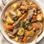 Creamy beef stew with tender beef, potatoes, carrots, mushrooms and peas served in a white bowl with a silver spoon