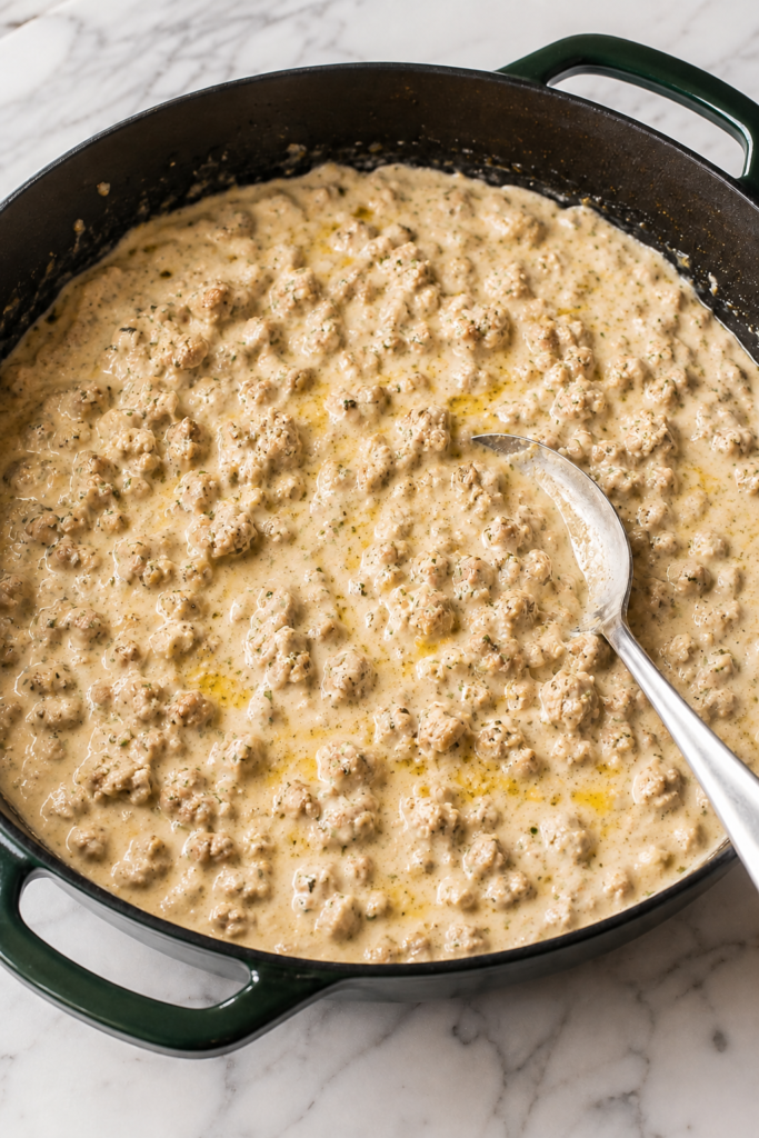 Creamy cowboy butter ground chicken sauce cooking in a dark green enamel skillet with garlic, herbs, lemon zest, and a silver spoon