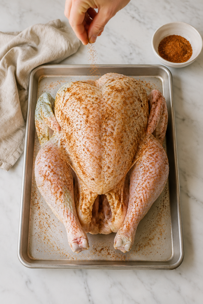Hand sprinkling Cajun dry-brine seasoning over a whole raw turkey on a rimmed sheet pan.