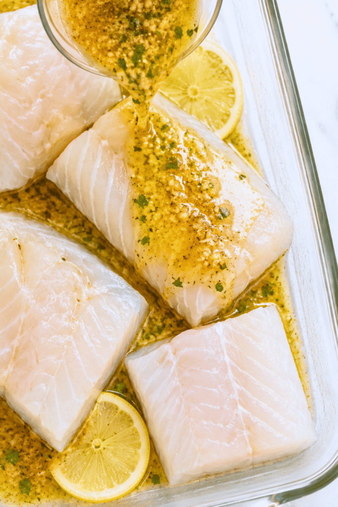 Lemon garlic butter mixture being poured over four raw cod fillets in a clear glass baking dish.