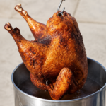 Crispy deep-fried Thanksgiving turkey being carefully lifted from a stainless-steel outdoor turkey fryer.