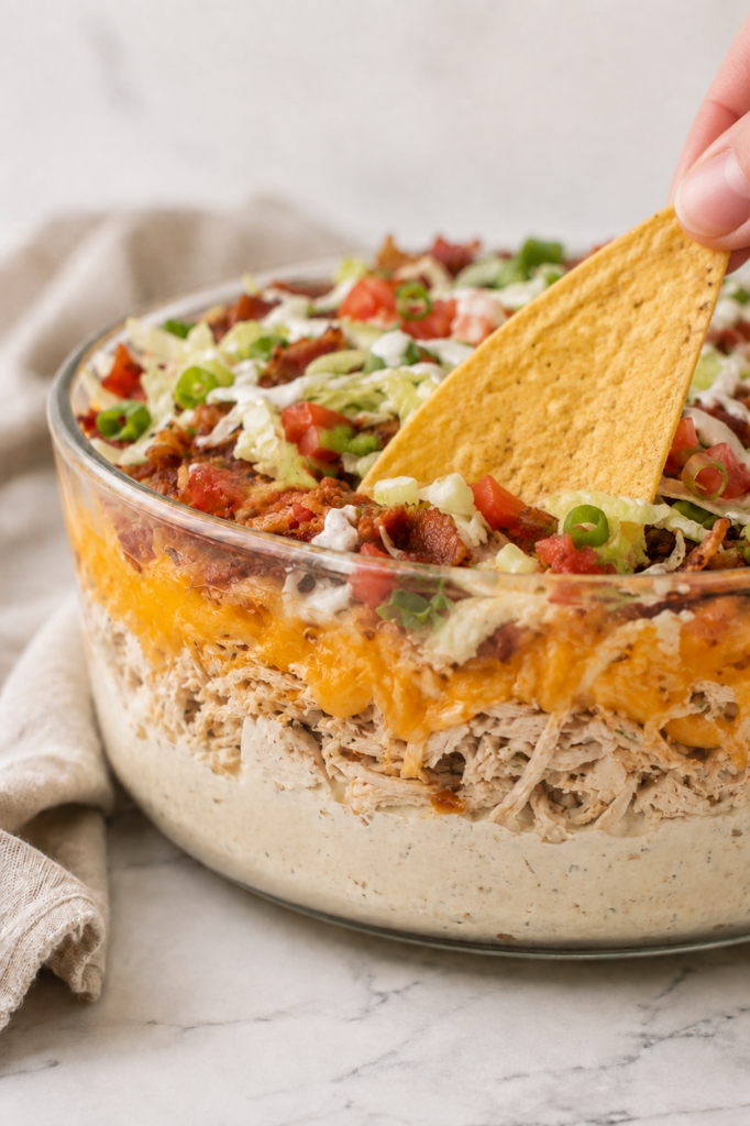 Tortilla chip dipping into five layer loaded crack chicken dip topped with bacon, lettuce, tomatoes, green onions, and ranch.