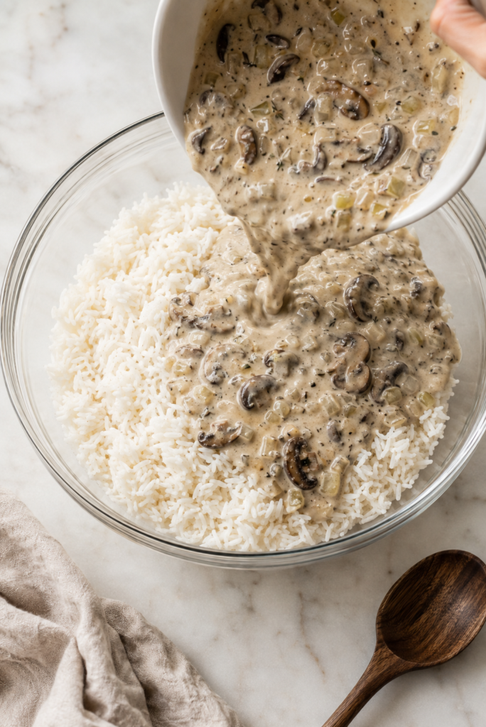 Creamy mushroom sauce with sliced mushrooms, onion, and celery being poured over cooked white rice in a glass mixing bowl.