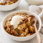 Warm pumpkin pecan cobbler with gooey caramel sauce, chopped pecans and melting vanilla ice cream in a white bowl.