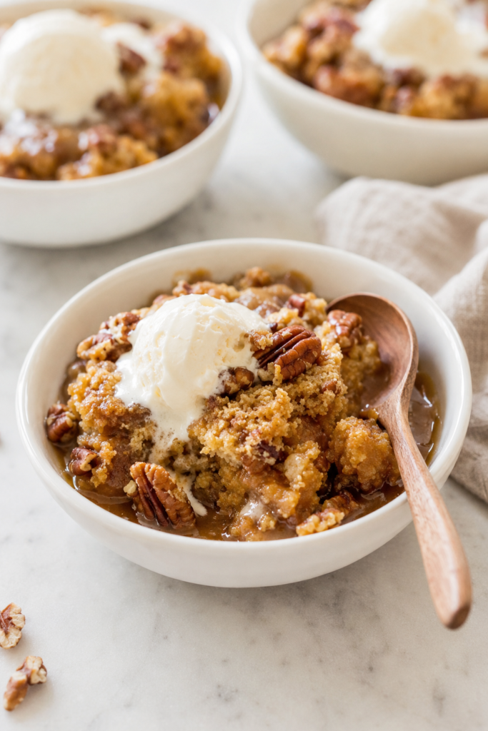 Warm pumpkin pecan cobbler with gooey caramel sauce, chopped pecans and melting vanilla ice cream in a white bowl.