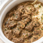 Tender slow cooker creamy beef tips with mushrooms and rich gravy served beside mashed potatoes in a white Crockpot.